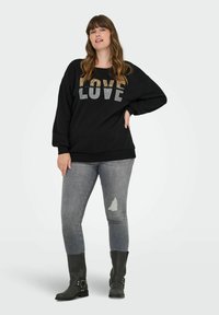 Sieviete, tērpusies melnā džemperī ar tekstu "MĪLESTĪBA", pelēkā plēstās skinny džinsās un melnās pusgarās zābaku, stāv ar vienu roku uz gurniem.