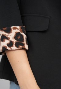 Svart blazer med leopardmønstret mansjett, som viser en jevn tekstur og en strukturert design med en frontlomme.
