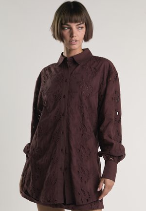 Frau mit kinnlangem braunem Haar, die eine lockere, langärmelige, dunkelbraune Bluse mit Knöpfen und floraler Ausschnitt-Stickerei trägt.