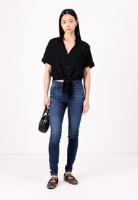 Cămașă neagră cu mâneci scurte și nasturi, legată în față, asociată cu blugi skinny de culoare albastru închis și pantofi tip balerin cu imprimeu leopard. Poartă o geantă neagră în mână.