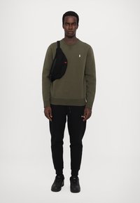 Olivengrønn hettegenser med en liten hvit logo, sammen med svarte joggebukser og svarte joggesko. En svart crossover-veske med røde detaljer brukes.