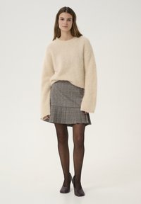 Licht beige fluffy trui met lange mouwen, gecombineerd met een geruite plooienrok in grijze en bruine tinten. Zwarte ondoorzichtige maillots en paarse hakken.