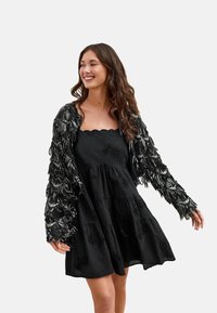 Zwarte jurk met geborduurde details, gelaagde ruches en een afgeronde halslijn, gecombineerd met een zwarte cardigan met franjes en zilveren kralen.