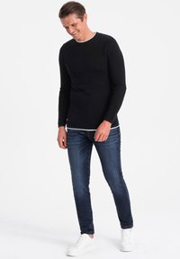 Black long-sleeve shirt with a white collar detail, paired with slim-fit dark blue jeans and white sneakers, standing on a plain background.