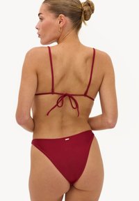 Femme avec les cheveux en chignon portant un bikini rouge avec de fines bretelles nouées dans le dos, vue de dos sur un fond blanc uni.