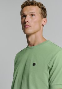 Groene katoenen t-shirt met ronde hals, voorzien van een klein zwart embleem op de borst. Glad aanvoelende textuur en een relaxte pasvorm.