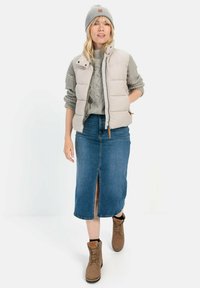 Beige Steppweste über einem grauen Strickpullover, kombiniert mit einem Jeansrock mit einem Schlitz vorne. Braune Schnürstiefel runden das Outfit ab.