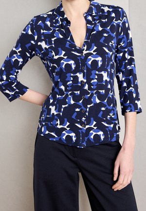 Vrouw draagt een blauwe, zwarte en witte blouse met knopen, driekwart mouwen en zwarte broek, staand met één hand op de heup.