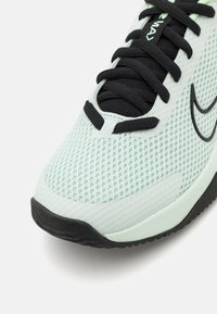 Nærbillede af en lysegrøn sportsko med sorte snørebånd, mesh-overdel, sort Nike swoosh og sort sålkanter.