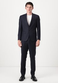 Jeune homme debout, portant un costume sombre à fines rayures avec un t-shirt blanc et des chaussures habillées noires, sur un fond blanc uni.