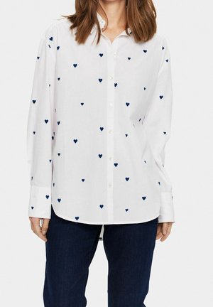 Frau trägt ein weißes Hemd mit kleinen marineblauen Herzmustern und dunkelblaue Jeans vor einem schlichten Hintergrund.