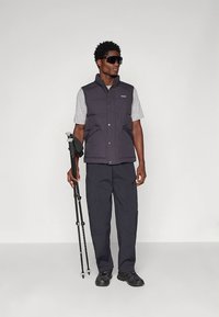 Tamsiai mėlyna paminkštinta liemenė dėvima ant pilkos marškinėlių, juodos kelnės ir vaikščiojimo lazdos. Tamsūs saulės akiniai papildo lauko drabužių derinį.