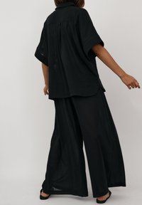 Osoba ubrana w luźną czarną koszulę z krótkim rękawem i szerokie spodnie, pozująca z wyciągniętymi ramionami na jednolitym jasnym tle.