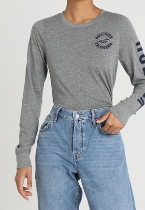 Person iført en grå langærmet skjorte med teksten "Hollister California" og lyseblå højtaljede jeans, hånd i lommen.