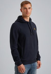 Sudadera azul marino hecha de tela suave, con capucha ajustable, bolsillo tipo canguro y pequeño parche de logo en el pecho.