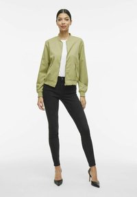 Giacca bomber verde chiaro con collo alto, chiusura con zip e polsini a costine, abbinata a jeans skinny neri e scarpe con tacco nere.