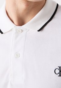 Detailní záběr na bílé polo tričko s černým lemem na límci a dvěma bílými knoflíky zapnutými na krku.