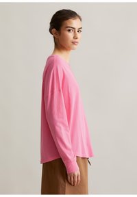 Frau mit zurückgebundenen dunklen Haaren trägt ein lockeres rosa Langarmshirt und einen braunen Rock, steht seitlich vor einem hellen Hintergrund.