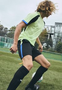 Heled roheline lühikeste varrukate spordisärk mustade elementidega, koos mustade lühikesed püksidega, millel on sinised küljeribad, mustad põlvekõrgused sokid ja mustad kingad.
