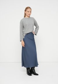Pulover gri tricotat cu mâneci lungi, butoane pe lateral; fustă maxi albastră dintr-un material neted; cizme negre cu toc, design minimalist.