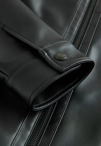 Veste en cuir noir avec fermeture éclair, matériau texturé, poignet avec un bouton-pression en métal et coutures contrastées.