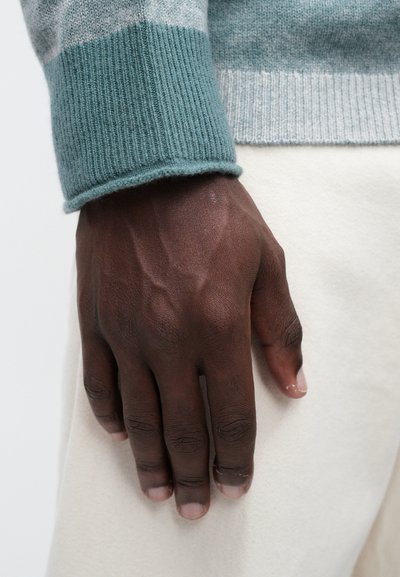 Heledu rohelise ribaga kampsuni varruka osa, mis katab osaliselt tumeda käe, millel on nähtavad veenid ja tugevalt tekstureeritud nahk, kreemja tausta ees.