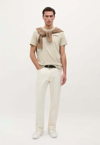 Beige T-Shirt mit Logo, kombiniert mit elfenbeinfarbener Hose und weißen Sneakers. Braun strukturierter Pullover über die Schultern gelegt, neutraler Hintergrund.
