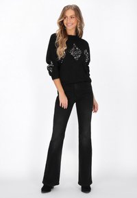 Schwarzer Pullover mit funkelnden, diamantmusterartigen Verzierungen, kombiniert mit schwarzen weit geschnittenen Jeans und schwarzen Stiefeletten. Glatte, gestrickte Textur.