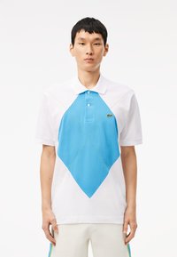 Camisa polo de algodón blanco con un gran patrón de diamantes azules. Presenta un cuello tipo polo, tres botones y detalles de costura en verde.