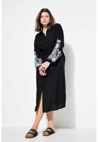 Schwarzes Wickelkleid mit langen Ärmeln, das mit weißer Blumenstickerei an den Manschetten verziert ist. Leichter Stoff, mit Schlitz vorne und einer gebundenen Taillenmaßnahme.