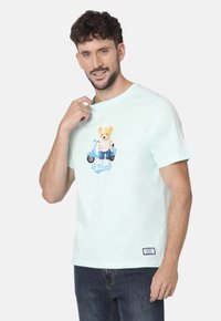 Camiseta de algodón de color verde menta claro con un gráfico de un oso de dibujo animado en patinete, mangas cortas y una pequeña etiqueta de marca en el dobladillo.