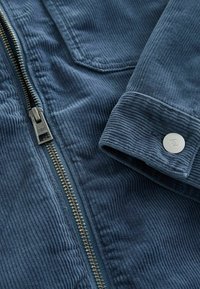 Veste en velours côtelé bleu foncé avec une fermeture éclair métallique visible et un bouton de manchette portant un logo gravé discret.