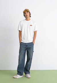 Hvid kortærmet t-shirt med en lille grafik, mørkeblå løse jeans, beige kasket og hvide sneakers med brune accenter.