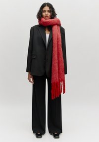 Zwarte op maat gemaakte blazer, witte blouse, brede zwarte broek en een lange, textuurijke rode sjaal met franjes, gedragen in een staande pose.