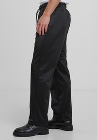 Sidevinkel af en person iført løse sorte bukser med elastisk talje og sorte loafers, stående på et gråt gulv mod en ensfarvet baggrund.