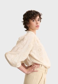 Crèmekleurige, geborduurde blouse met ronde en vierkante oogjespatronen; lichtgewicht stof, verlaagde schouders en gepofte mouwen.