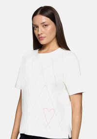 Hvid kortærmet skjorte med et diamantskema af iriserende perler og en pink hjerte-detalje nær bunden. Glat, letvægtsstof.