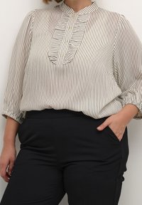 Blouse rayée blanche et noire avec un col haut, un devant plissé et des boutons. Tissu doux et léger, associé à un pantalon noir.