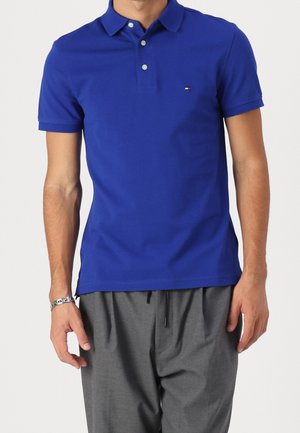 Mann mit einem taillierten royalblauen Polohemd mit kurzen Ärmeln und Knöpfen, grauen Jogginghosen mit Kordelzug und einem silbernen Armband am linken Handgelenk.