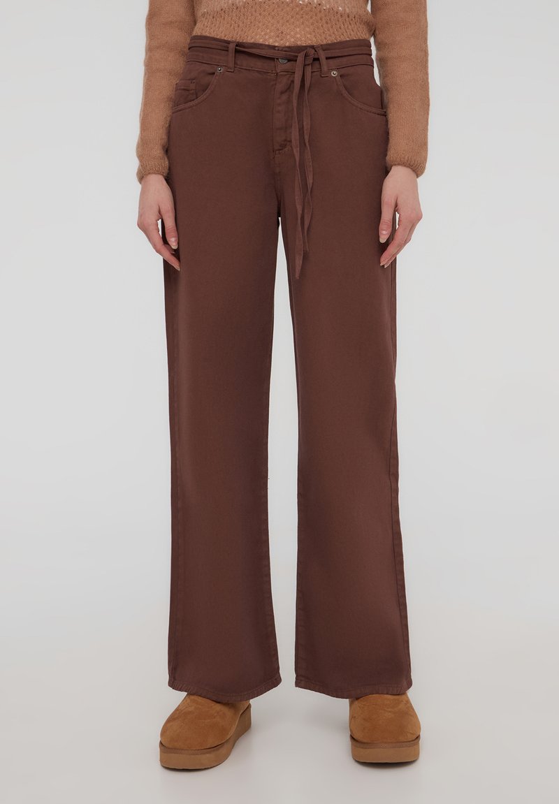 Pantaloni larghi marroni realizzati in tessuto liscio, con una vita regolabile legata e tasche laterali, abbinati a zoccoli marroni chiari.