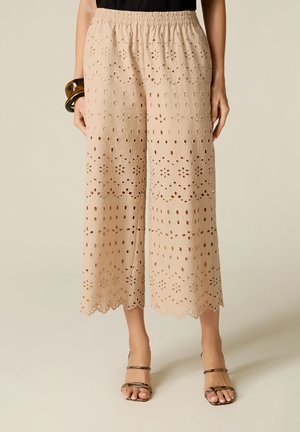 Beige wijde cropped broek met bloemvormige opengewerkte details, gegolfde zoom, elastische tailleband, gedragen met open schoenen en een zwart topje.