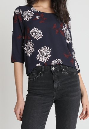 Frau trägt eine marineblaue Bluse mit weißen und roten Blumenmustern und schwarze hoch taillierte Jeans, stehend vor einfachem Hintergrund.