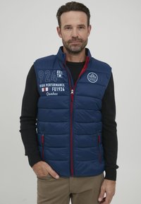 Temno modra puhaste jakna brez rokavov z rdečimi detajli in zadrgami. Ima kvadratni dizajn, vezenine logotipov in stranske žepe. Narejena je za nošenje čez črno pulover.