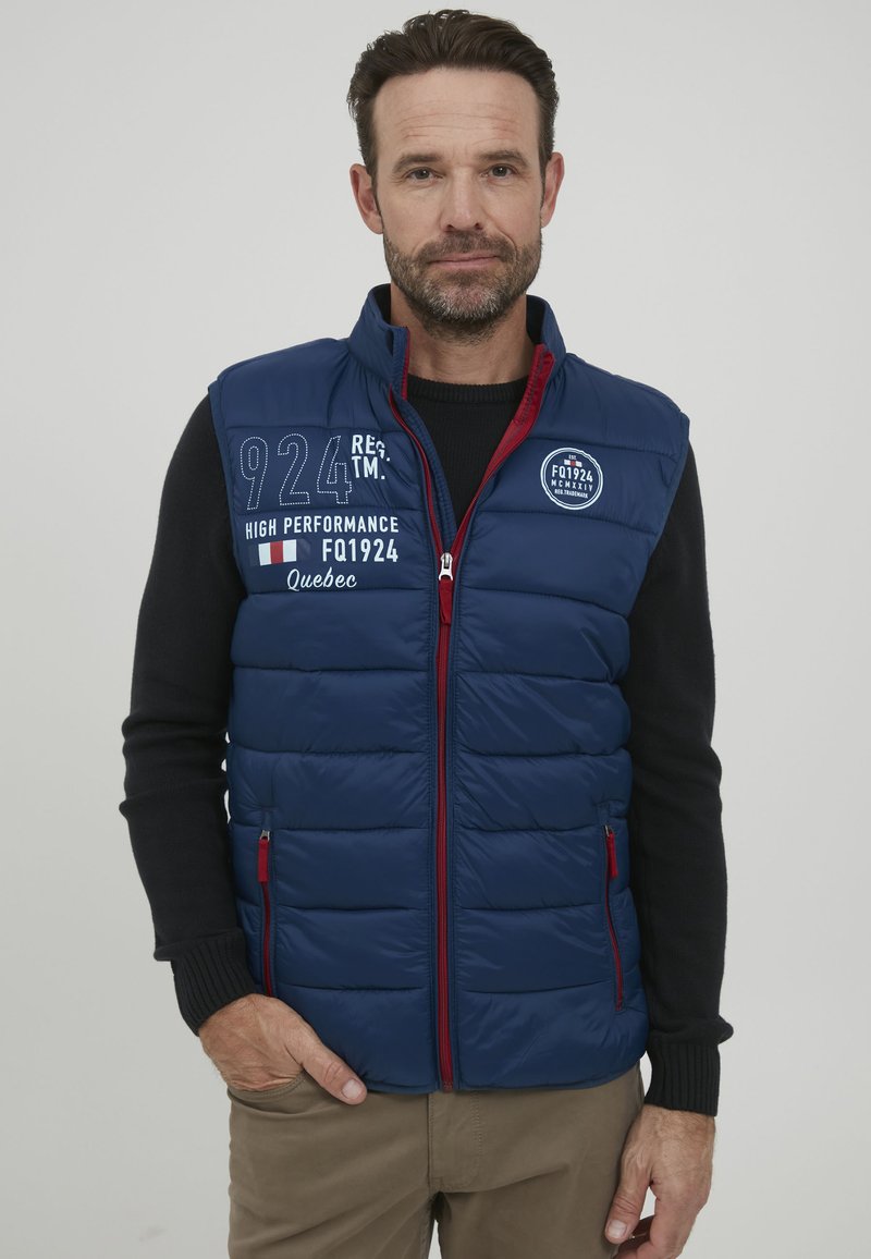 Temno modra puhaste jakna brez rokavov z rdečimi detajli in zadrgami. Ima kvadratni dizajn, vezenine logotipov in stranske žepe. Narejena je za nošenje čez črno pulover.