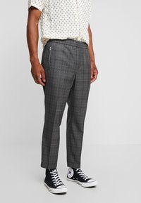 Man med grå rutiga capribyxor med dragkedjefickor på sidan, vit mönstrad kortärmad skjorta och svarta högskurna sneakers.