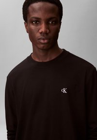 Junger Mann mit kurzen lockigen Haaren, der ein schwarzes Langarmshirt mit weißem "cK"-Logo vorne trägt, vor einem einfarbigen grauen Hintergrund.