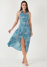 Un vestito senza maniche, con bottoni, realizzato in tessuto teal con motivi di foglie bianche, dalla silhouette fluida e con uno spacco laterale per facilitare i movimenti.