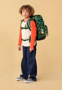 Groene rugzak met een webpatroon, oranje shirt met lange mouwen, blauwe jeans en zwart-witte geblokte schoenen. Kind dat tegen een neutrale achtergrond staat.