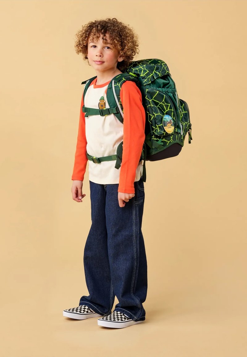 Groene rugzak met een webpatroon, oranje shirt met lange mouwen, blauwe jeans en zwart-witte geblokte schoenen. Kind dat tegen een neutrale achtergrond staat.