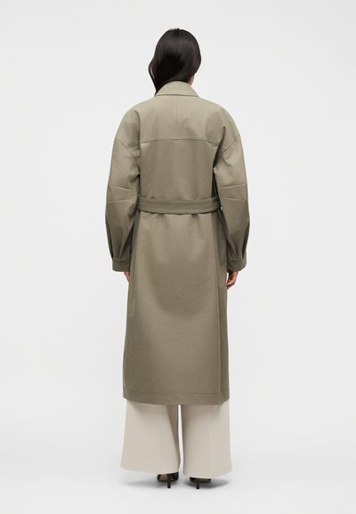 Γυναίκα με μακριά σκούρα μαλλιά, που φοράει ένα χακί καμπαρντίνα με ζώνη πάνω από φαρδιά κρεμ παντελόνια και μαύρες γόβες, όρθια μπροστά σε λευκό φόντο.